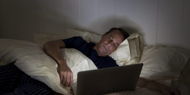 A man lies in bed in a dimly lit room, propped up by pillows, looking intently at a laptop. He is covered with a blanket and appears to be either working or watching something on the screen.