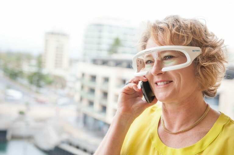 A woman with blonde curly hair is wearing white smart glasses and a yellow top. She is smiling while talking on a mobile phone, her re-timer glasses contributing to her vibrant energy. The background shows a blurred urban landscape with buildings and trees.