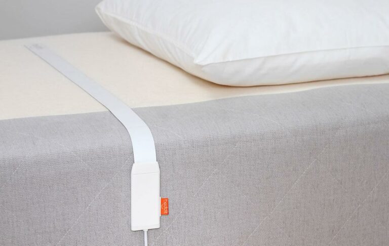 A white sleep tracker is placed on a bed. The sensor strip is positioned under the bedsheet, with the attached white monitor resting on the edge of the mattress. A white pillow is situated at the head of the bed. The tracker, often praised in Beddit reviews, has a small orange tag.