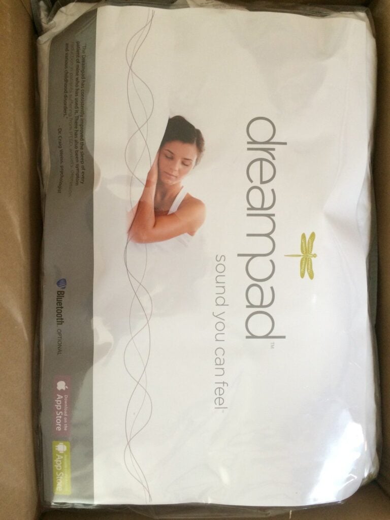 A packaged "dreampad" with a logo featuring a dragonfly and the tagline "sound you can feel!" showcases a person resting their head on a pillow. The packaging highlights Bluetooth and iOS app compatibility, making Dreampad an essential sleep accessory.