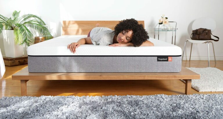 A woman with curly hair is lying on her side on a gray YogaBed mattress in a bright, minimalist bedroom. The room features a wooden bed frame, a potted plant, a white chair, a gray shag rug, and a brown bag on a table.