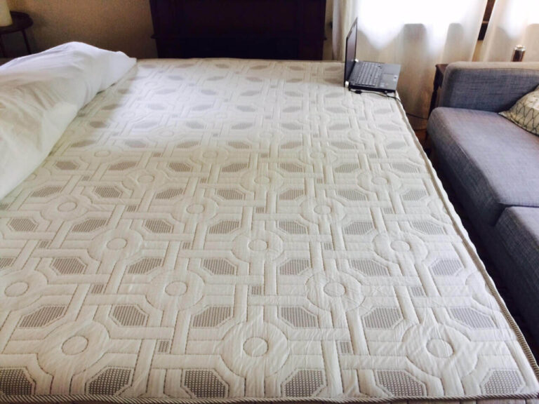 A large quilted 4Sleep Mattress with a geometric pattern on a bed, partially covered by pillows on one end. On the other end, there is an open laptop computer resting near the edge. The room's background includes a couch and some drapery-covered windows.