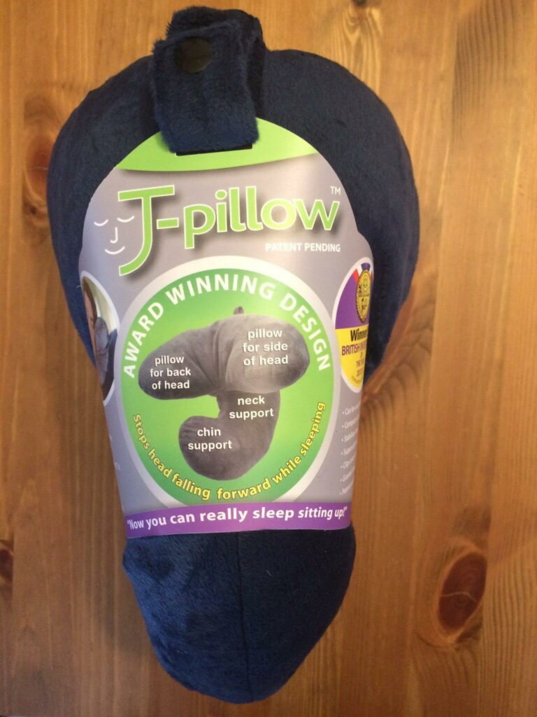 A J-Pillow in navy blue rests on a wooden surface, showcasing its unique design that supports the back of the head, side of the head, neck, and chin. The packaging highlights its award-winning design with the tagline, "Now you can really sleep sitting up," making it clear why the J Pillow stands out.