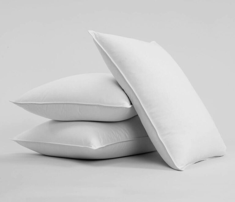 Three Brooklinen Pillows are stacked against a plain gray background. Two pillows are laid flat on the bottom, and the third pillow is balanced upright on top of them.