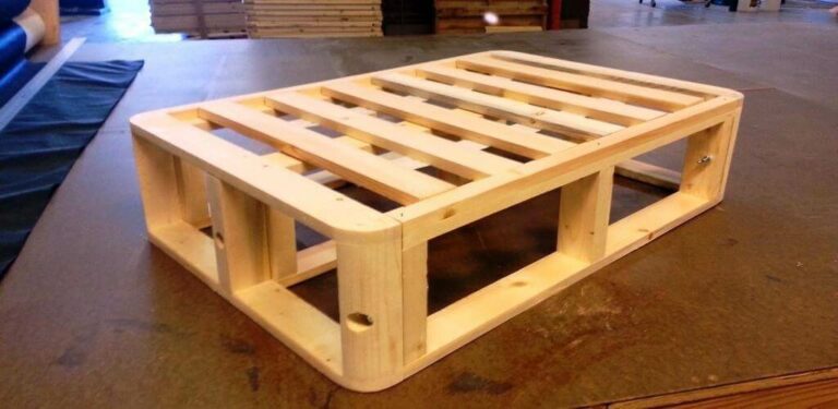 A neatly crafted wooden pallet with smooth, rounded edges and cut-outs for forklift access sits on a flat surface in a workshop. Among the background's wooden materials and workspace elements, it's easy to imagine this sturdy pallet repurposed into a custom bed frame.