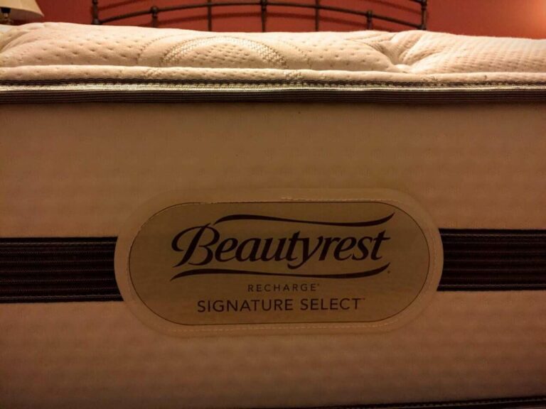 A close-up view of the side of a mattress with the label "BeautyRest Recharge Signature Select." The label is affixed to a beige and white textured fabric with a brown stripe running horizontally. A dark metal bedhead is visible in the background.