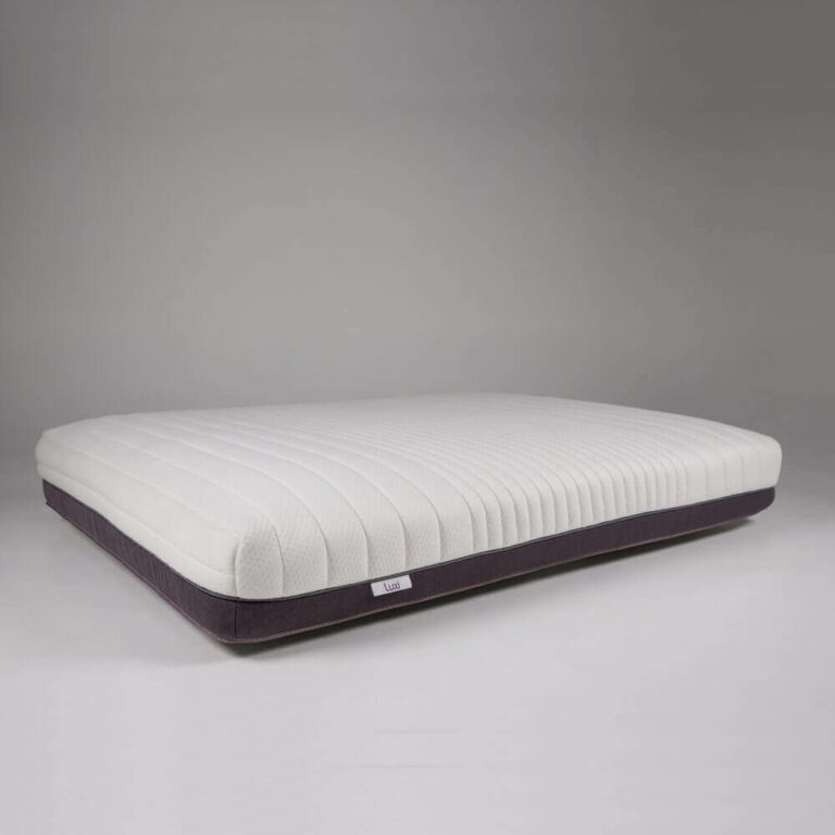 A Luxi Mattress lies flat on a plain, light gray background. The mattress has a white, textured top with subtle stripes and a dark gray base. A small white tag with text is visible on the side.