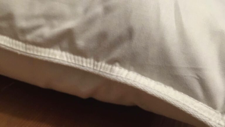 Close-up photo of a Ritz Carlton pillow in a white pillowcase, resting on a wooden surface. The pillowcase shows some wrinkles and stitching along the edge, highlighting its luxurious detail.