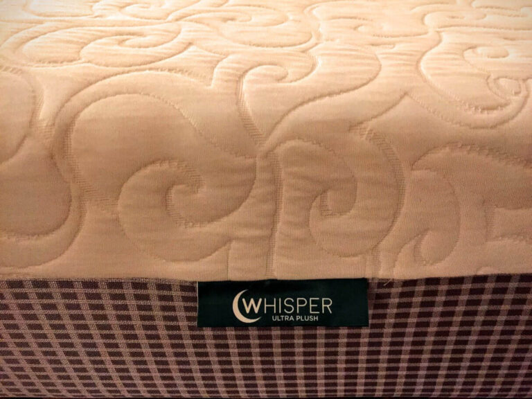 Close-up of a mattress with an intricate, swirling quilted pattern on its beige surface. The label on the side reads "Whisper Bed Ultra Plush" against a dark green background. The mattress sides have a checkered fabric pattern in shades of brown and white.