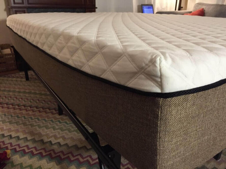 A close-up view of a bed with a quilted white mattress on top of a textured, Loblolly pine brown bed frame. The bed is set on a multicolored zigzag-patterned rug, and a laptop is visible in the background on a table.