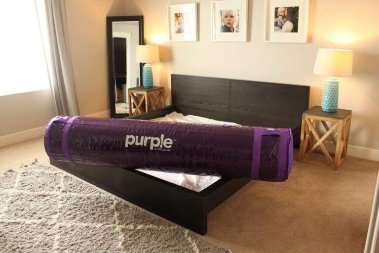 A bedroom with a beige carpet and walls contains a bed frame without a mattress. A rolled-up Purple Mattress is placed on the bed frame, its cover bearing the "Purple" label. Twin nightstands with lamps and framed photos hang above the bed, and a large mirror stands in a corner.