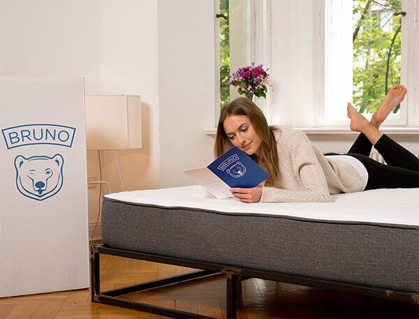 A woman lies on her stomach on a bed, reading a Bruno booklet. The mattress is set up in a bright room with a window, a nightstand, and a lamp. A Bruno-branded box with a bear logo is placed next to the bed. A vase with flowers is visible on the nightstand.