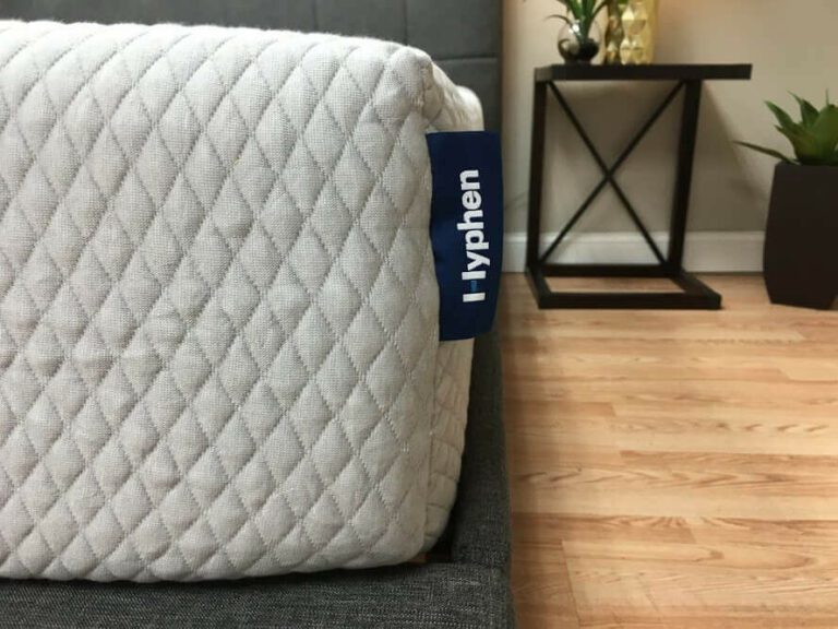 Close-up of a corner of a Hyphen Mattress with a white quilted cover, showing a blue fabric tag labeled "Hyphen." The mattress is on a grey bed frame, and the room has a light wooden floor. In the background, there is a black side table with a small plant on top and another plant on the floor.