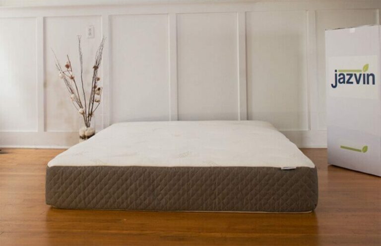 A mattress sits on a wooden floor in front of a white-paneled wall. To the right, there is a tall, white box with the brand name "Jazvin" printed on it in blue and green letters. A decorative vase with branches stands to the left of the mattress.