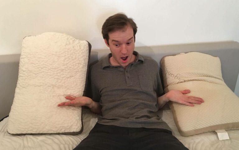 A person with a surprised expression sits on a bed between two pillows. They are spreading their arms wide open towards the pillows, which are propped up on either side of them. The individual is wearing a gray polo shirt.