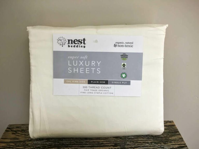 A package of Nest Bedding luxury sheets is displayed. The label indicates it is a California King size with a plain hem, 300 thread count, made from fair trade organic cotton. The sheets are noted to be organic, natural, and non-toxic. The color of the sheets is off-white.