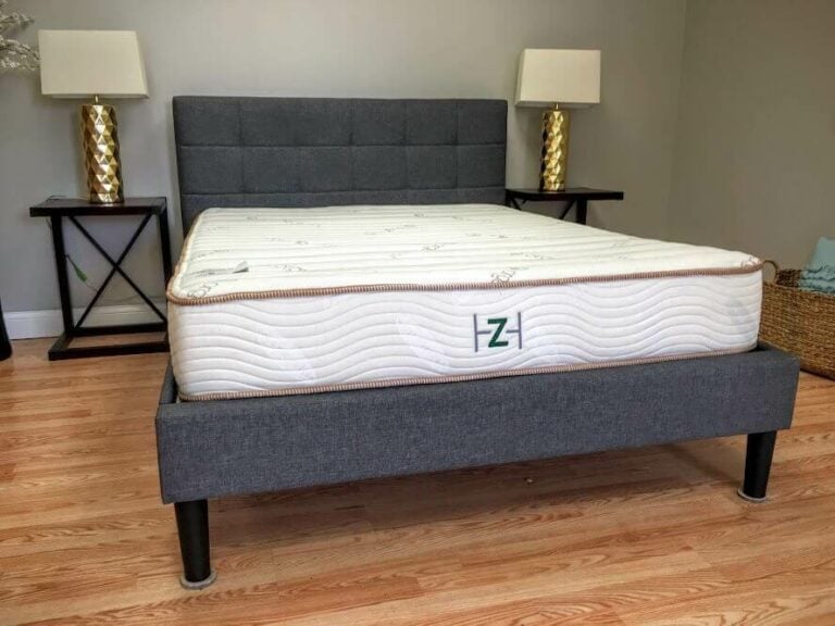 A neatly made bed with a gray upholstered headboard and frame beckons tranquility. The white mattress, edged in light beige, features a green 'Z' logo from Zen Haven. Two nightstands flank the bed, each holding a lamp with a gold base and cream-colored shade. The wooden floor completes the serene setup.