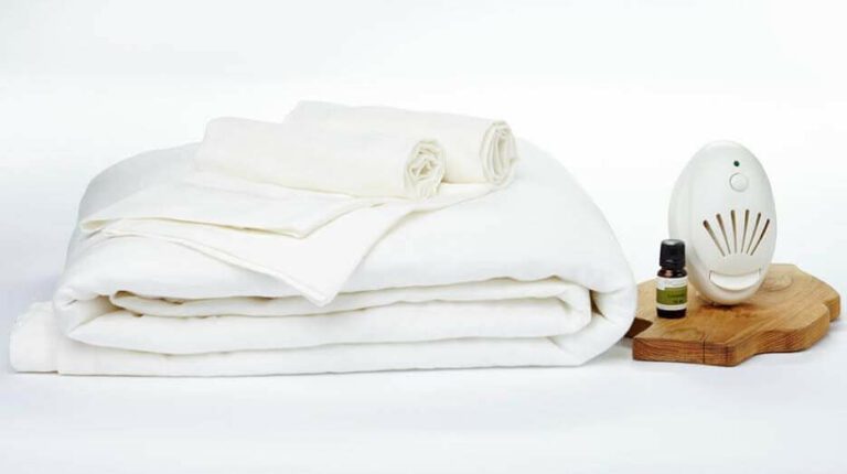 A neatly folded white blanket and white towels are placed on a white surface. Next to them is a wooden platform holding a small white electric diffuser and a small bottle of lavender essential oil for sleep. The scene has a clean and organized appearance.