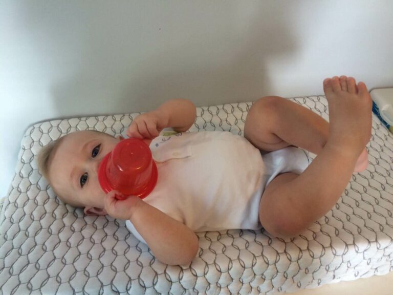 A baby in a white onesie lies on a patterned changing pad, holding and sucking on a red toy or cup. With one leg bent and the other extended, the wide-eyed infant gazes up at the camera. The background is a plain wall.