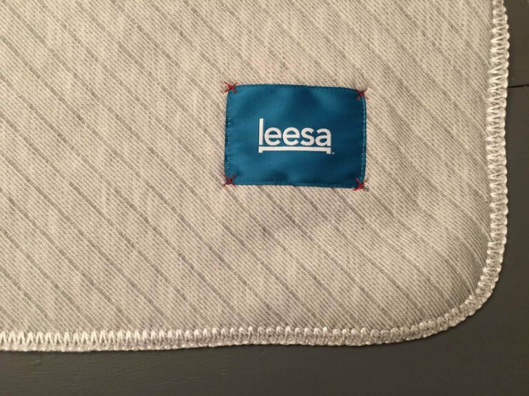 Close-up of a gray fabric with a textured pattern and rounded edges, featuring a blue rectangular label with the white text "leesa" in the center. The leesa blanket's label is stitched on with red thread at the corners.