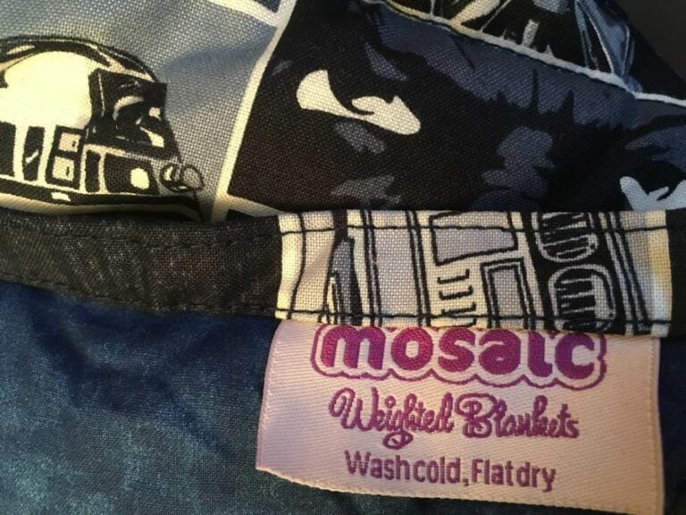 Close-up of a weighted blanket with a "Mosaic Weighted Blankets" label in pink and white, noting care instructions to "Wash cold, Flat dry." The blanket features a pattern with Star Wars-themed characters and vehicles in black and white.