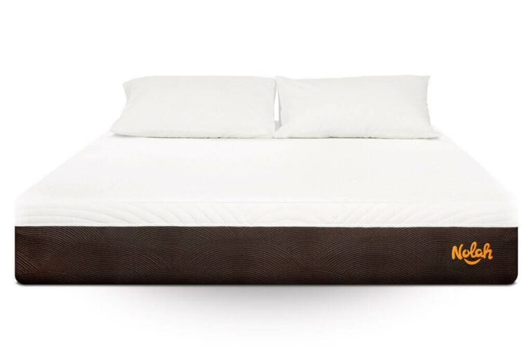 A Nolah Mattress with a white mattress and two white pillows sits atop a brown base featuring a textured pattern. The brand name "Nolah" is visible in orange on the bottom right corner. The bedding appears clean and neatly arranged.