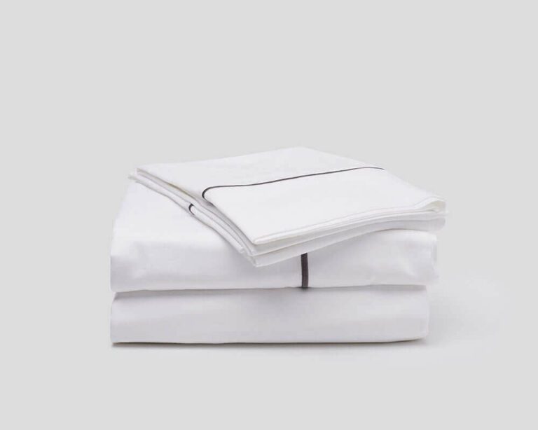 A neatly folded set of white bed sheets with a black trim on the pillowcases, stacked on top of each other against a plain grey background.