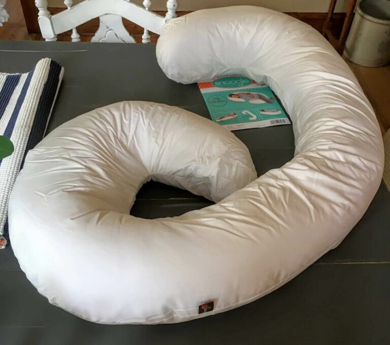 A large, white Snoogle C-shaped body pillow lies on a gray surface. Beside it, a packaged cover featuring an image of the pillow with a person using it, along with some text, is visible. The setting appears to be an indoor room with a white chair and other household items nearby.