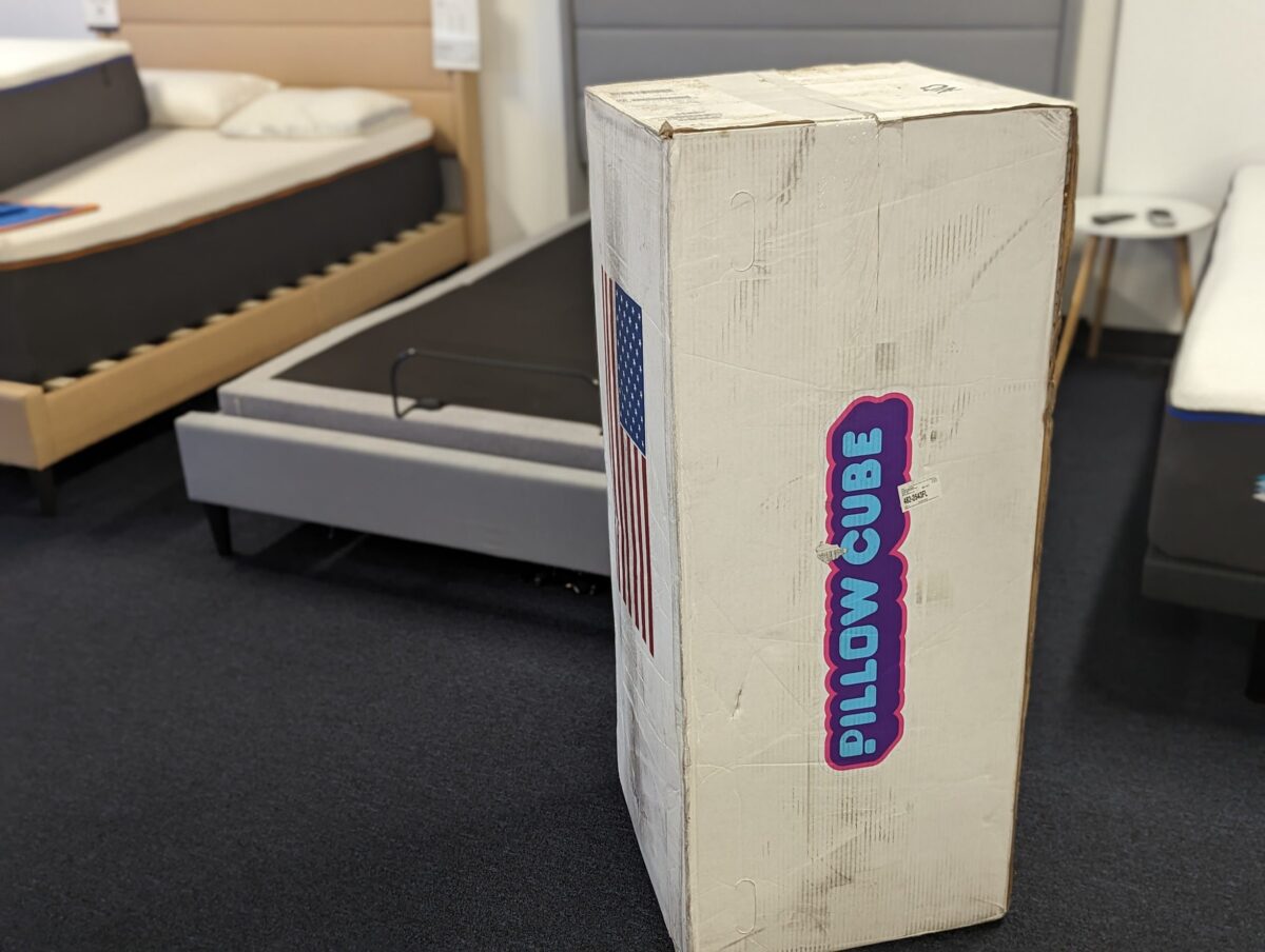 A large white rectangular box labeled "Pillow Cube" stands upright in the middle of a showroom with several mattresses and bed frames in the background. The box features an American flag graphic on one side.