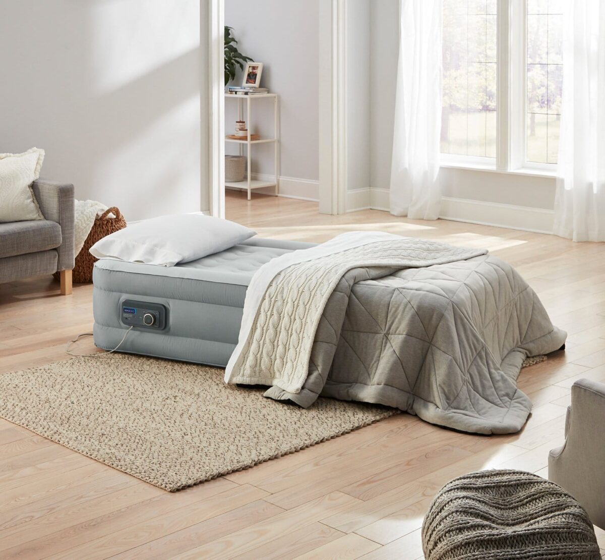 An air mattress with a pillow and blanket is set up in a bright, modern living room with large windows. A beige rug covers part of the wooden floor, and there’s a gray armchair and a small shelving unit with decor items in the background.