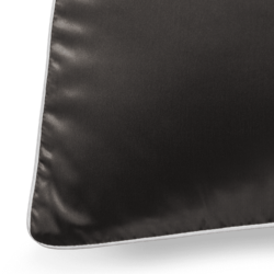 A close-up of a black silk pillowcase with white piping along the edges. The smooth texture of the pillowcase is highlighted by the light, creating a subtle sheen. Only the corner and a small portion of the front of the pillowcase are visible.
