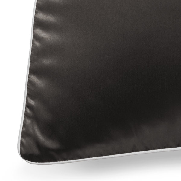 A close-up of a black silk pillowcase with white piping along the edges. The smooth texture of the pillowcase is highlighted by the light, creating a subtle sheen. Only the corner and a small portion of the front of the pillowcase are visible.