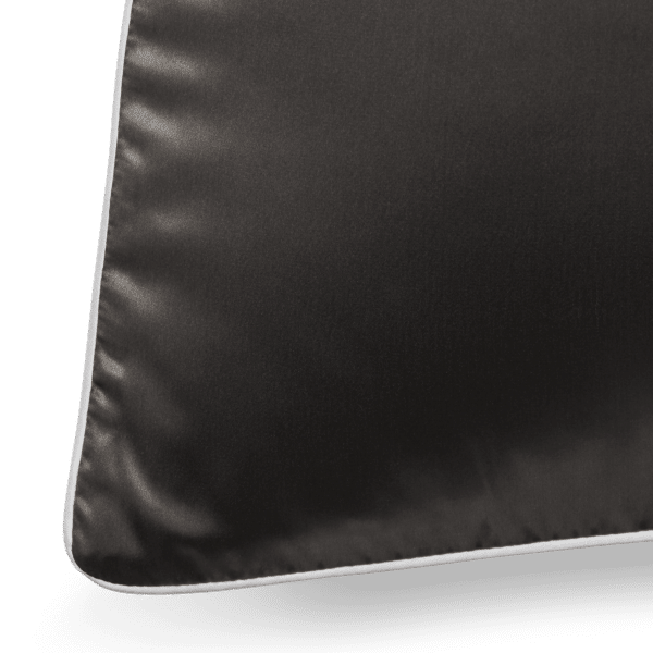 A close-up shot of a black Silk Pillowcase with elegant white piping along its edges. The fabric boasts a smooth and glossy texture, and the seam along the piping is clearly visible. The lighting accentuates the luxurious quality of the pillowcase's material.