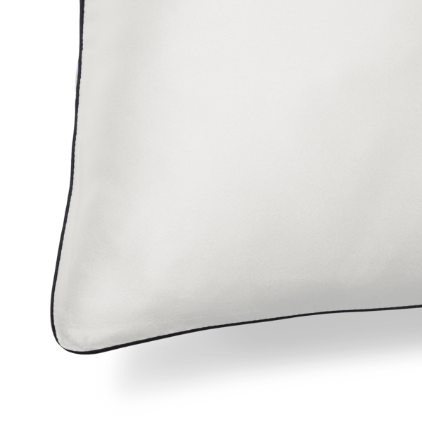 A close-up image of the Silk Pillowcase, showcasing its smooth white fabric with a dark border on one side. The pillowcase features a rounded corner, and the focus is on the texture and finish of the luxurious silk material, with the background in shadow.