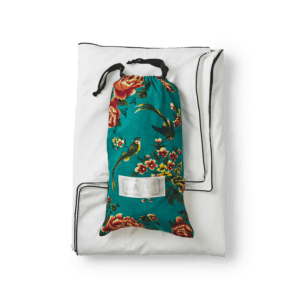A white pillow with black piping along the edges is laid flat. On top of it, there is a drawstring bag made of teal fabric adorned with a vibrant floral pattern featuring red and yellow flowers and green leaves. The bag, part of the Open Road Portable Silk Throw Set, has a clear label with text on it.