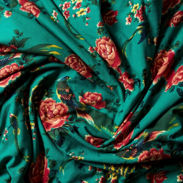 The Open Road Portable Silk Throw Set showcases a vibrant turquoise fabric adorned with intricate patterns of red and pink peonies, green leaves, and colorful birds. This elegant and lively design is softly draped to highlight the detailed floral and avian motifs.