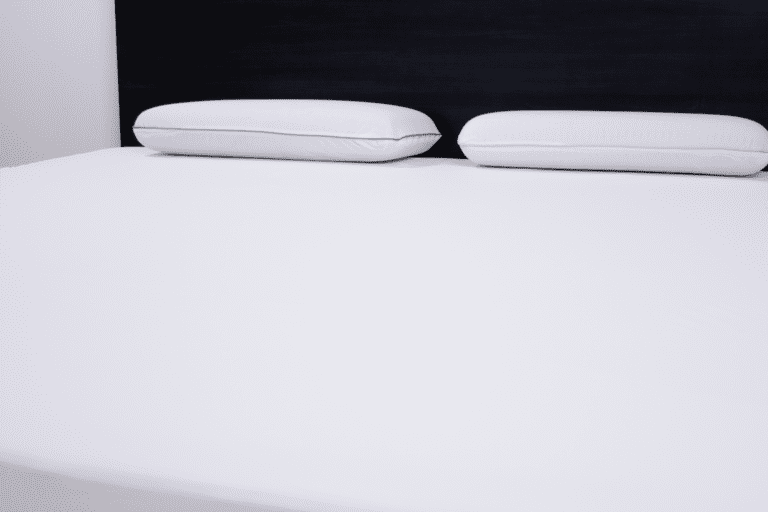 A neatly made bed with Solis Quiet Comfort™ featuring a white sheet and two white pillows is shown. The headboard is dark, providing a striking contrast to the light bedding. The room appears clean and minimalistic.