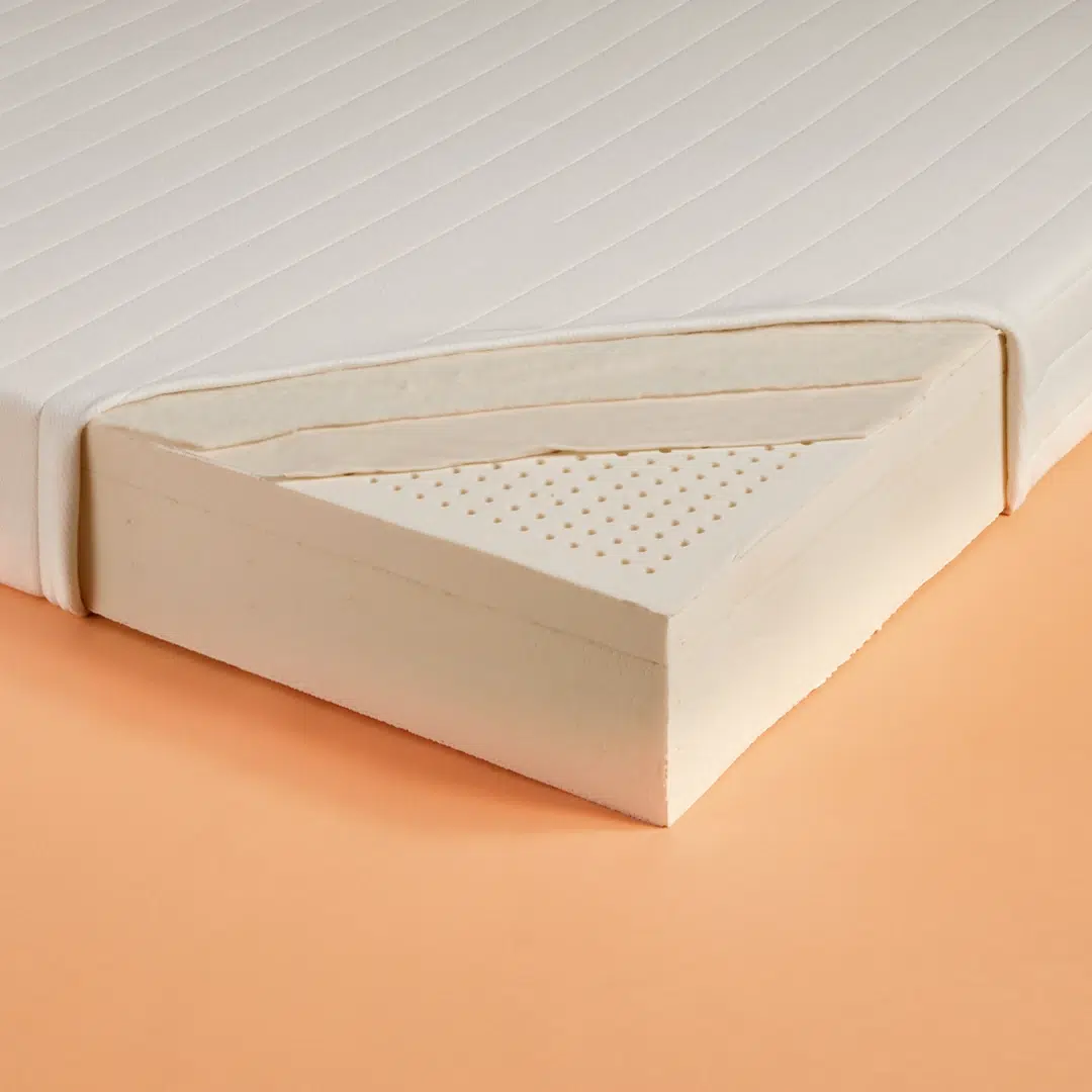 An image shows a mattress with one corner cut away to reveal its interior layers. The mattress has a white fabric cover, and underneath, there is a thick foam base layer topped with a perforated foam layer. The background is a smooth, light orange surface.