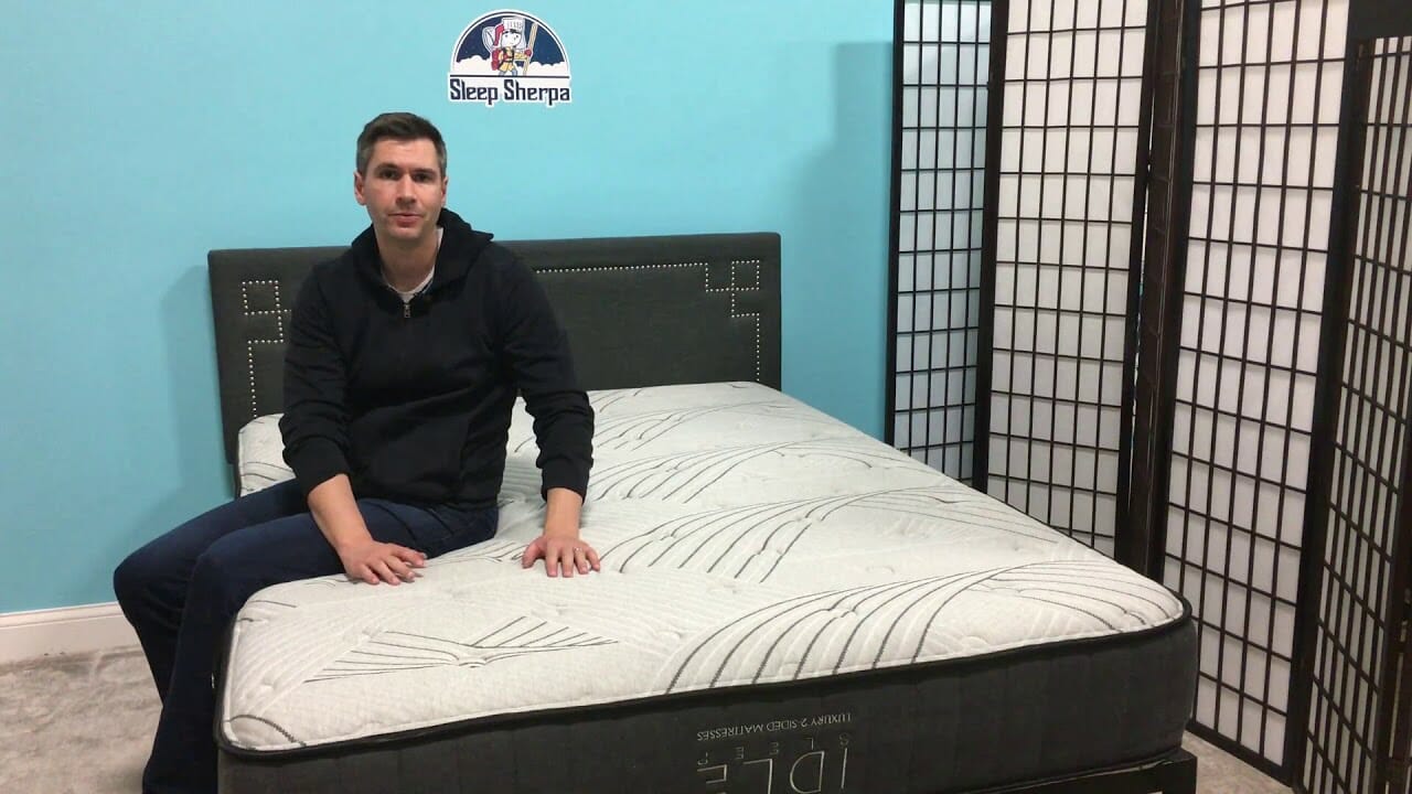 A person sits on the edge of a large bed in a room with light blue walls, idly adjusting their dark hoodie and jeans. A black headboard frames the bed, and a room divider with multiple panels stands in the background. The wall features a logo that reads "Sleep Sherpa.