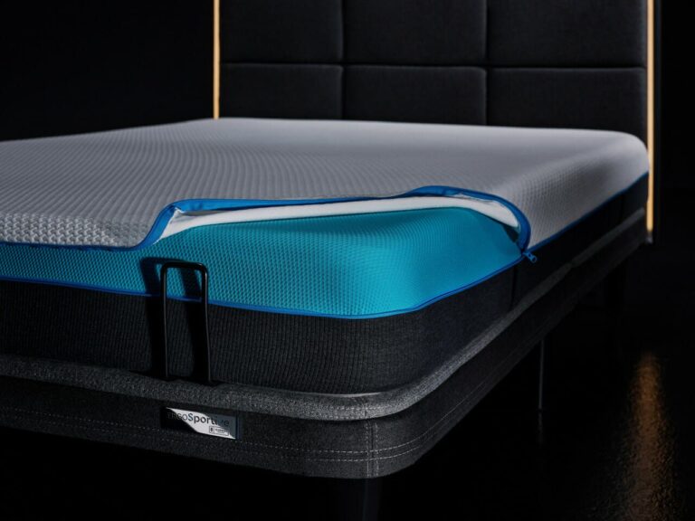 The Ergo Sportive Adjustable Smart Bed boasts a modern design with a dark fabric headboard and features a layered mattress. The top layer of the mattress is white with a blue zipper, which reveals blue and darker sublayers underneath. The bed’s frame is finished in dark gray, and the label "proSport" is prominently displayed on the side.