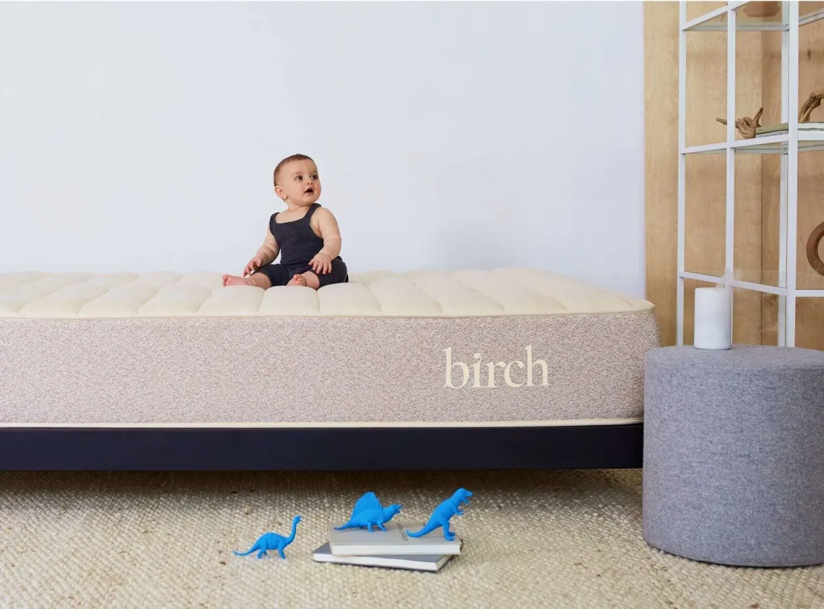 A baby wearing a dark tank top sits on a beige Birch Natural Mattress. The baby is looking to the left. In front of the mattress are three blue toy dinosaurs and a closed book. Beside the mattress is a gray cylindrical stool, and the background features a bookshelf.