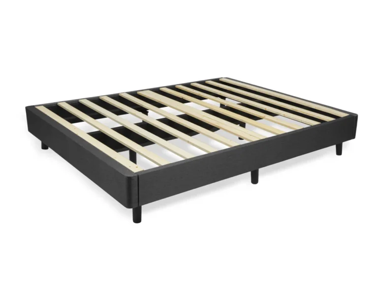 The "Foundation" is a minimalistic black bed frame with a wooden slat support system, shown from an angle. It features six sturdy black legs and lacks both headboard and footboard, resting directly on the floor. The design emphasizes simplicity and functionality.