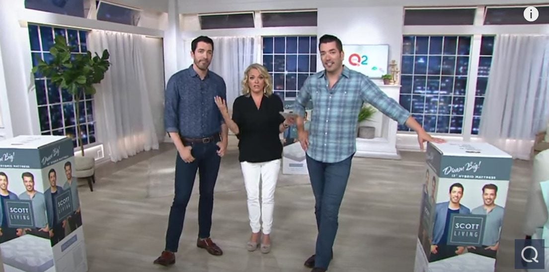 Three people are standing in a brightly lit studio with QVC2 branding in the background. Two men in casual shirts and jeans are posing with a woman dressed in a black shirt and white pants. They are surrounded by large product boxes, including one featuring the new QVC mattress with images of the men on it.