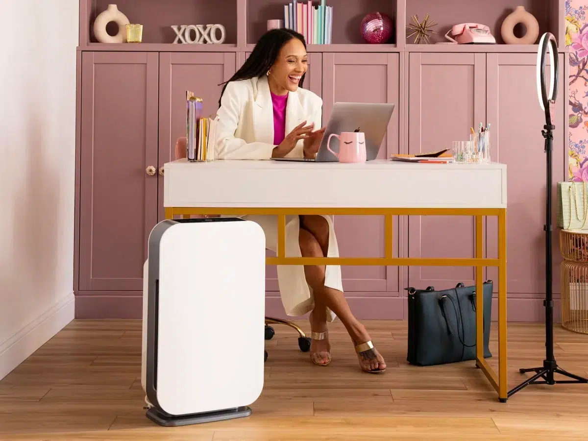 A person sits at a desk in a stylish, pink-themed home office, smiling at a laptop. Nearby are a ring light, office supplies, and an Alen air purifier. The background features a cabinet with decor, including a sign that reads "XOXO" and various decorative items.