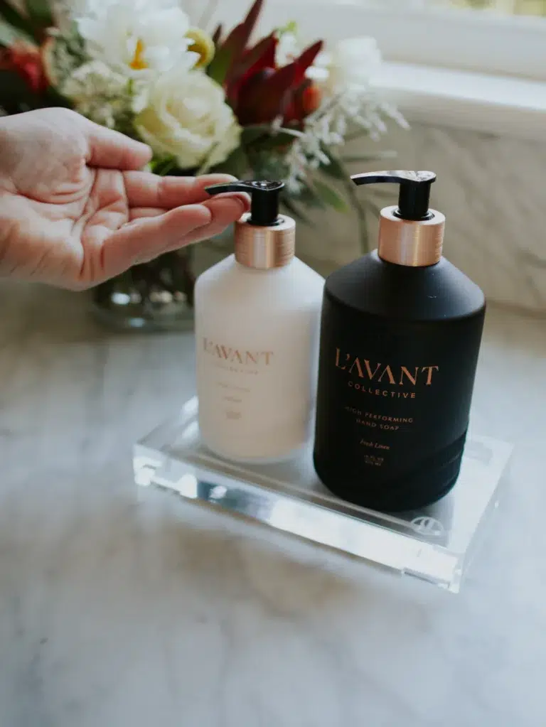 A hand pressing the pump of a white soap dispenser beside a black soap dispenser, both labeled "L’AVANT COLLECTIVE," positioned on a clear Lucite Tray - Medium. In the background, there is a floral arrangement on a marble countertop.