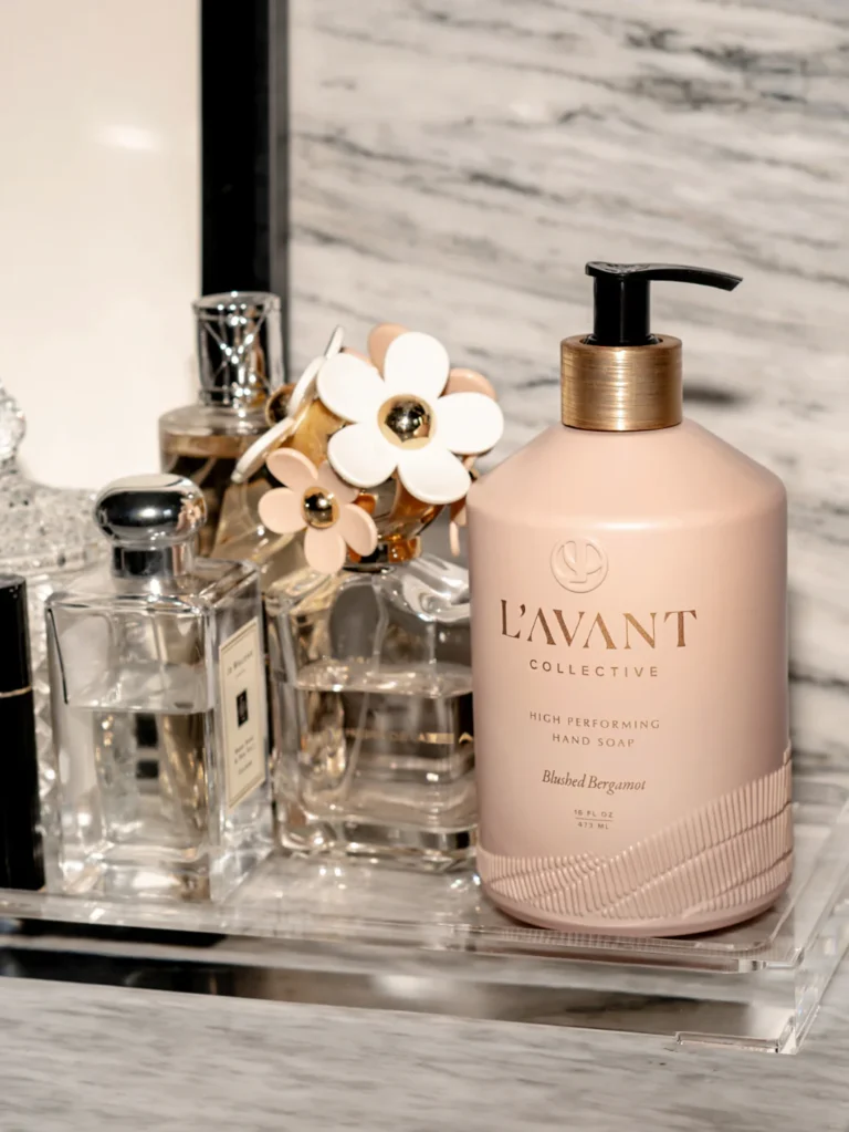 A bottle of "High Performing Hand Soap - Blushed Bergamot - L'AVANT Collective" sits on a counter next to several perfume bottles and decorative items. The background features a marble surface.