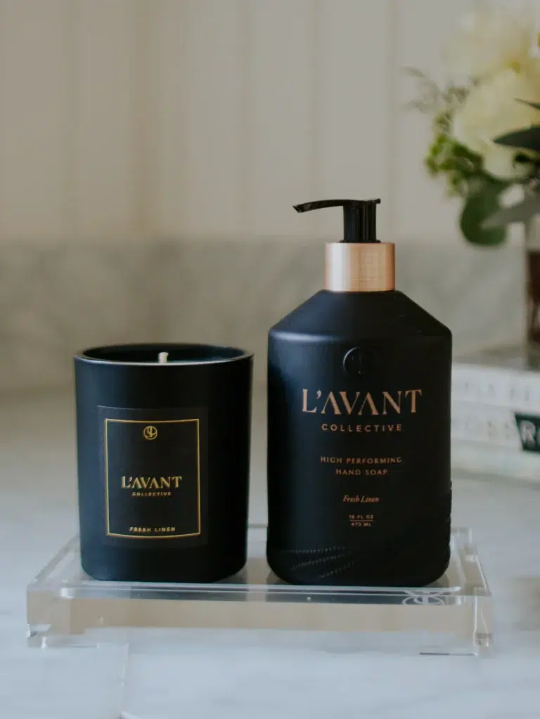 A black candle container and a black pump bottle of L'Avant Collective High Performing Hand Soap, both labeled "Fresh Linen," sit on a Lucite Tray - Medium from L'AVANT Collective. In the background, there are blurred flowers and a book with partially visible text.