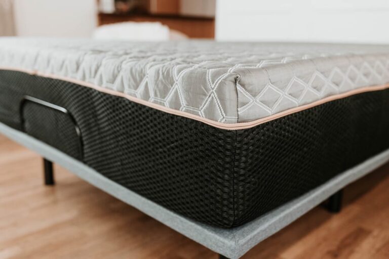 A close-up of a BedTech Copper Lux Hybrid 14" mattress featuring a diamond-patterned topper and a black textured base. The bed is positioned on a grey bed frame with visible legs, set in a room with wooden flooring and blurred furniture in the background.