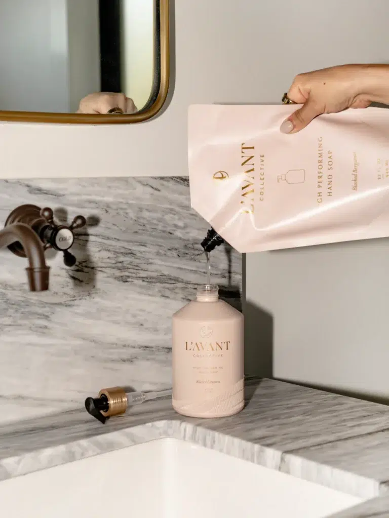 A person's hand is refilling a container of L'AVANT Collective High Performing Hand Soap in the Blushed Bergamot scent using a L'AVANT Collective refill pouch over a marble countertop. The scene includes a sink with a black faucet and part of a mirror.