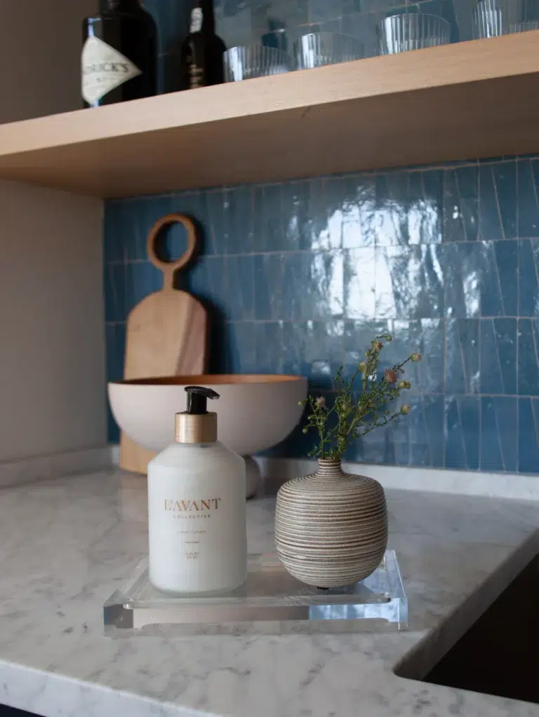 A kitchen countertop showcases a Lucite Tray - Medium from L'AVANT Collective, containing a bottle of Lavant hand soap and a small striped vase with flowers. Situated behind the tray are a wooden cutting board and a bowl, set against a blue tiled backsplash. Upper shelves display glassware and bottles.